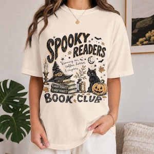 Könnte beinhalten: Ein cremefarbenes T-Shirt mit einem schwarzen Grafikdruck mit dem Text "SPOOKY READERS BOOK CLUB" und "Running on coffee, horror & chapters". Das Design zeigt einen Hexenhut auf Büchern, eine schwarze Katze, einen Kürbis und eine Kerze.