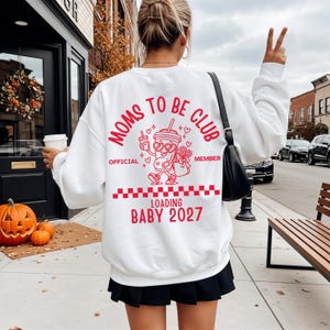 May include: A white long-sleeve sweatshirt featuring a red graphic on the back. The design includes the text "MOMS TO BE CLUB," "OFFICIAL MEMBER," and "LOADING BABY 2027," alongside a cartoon cup with heart-shaped sunglasses and a checkered pattern.