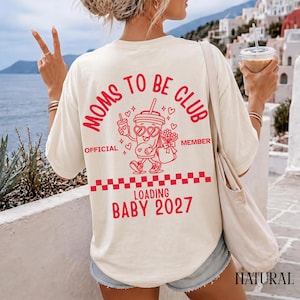 Può includere: Una t-shirt color crema con una grafica rossa sul retro. Il design include il testo MOMS TO BE CLUB, OFFICIAL MEMBER, LOADING BABY 2027, un motivo a scacchi e un personaggio a forma di tazza con occhiali a cuore che tiene dei fiori.