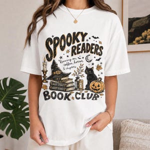 Könnte beinhalten: Ein weißes T-Shirt mit einem Grafikdruck, der den Text "SPOOKY READERS BOOK CLUB" und "Running on coffee, horror & chapters" zeigt. Das Design umfasst einen Hexenhut auf einem Bücherstapel, eine schwarze Katze, einen Kürbis und Herbstblätter.