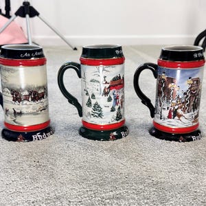 May include: Three ceramic Budweiser beer steins featuring winter scenes. Each stein has a dark green handle, a red rim and base with gold accents, and a cylindrical body decorated with detailed illustrations of snowy landscapes and horse-drawn carriages.