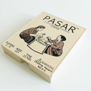 May include: A board game box titled "PASAR" with illustrations of people at a market. The box is beige with black text and illustrations. The game is for 1-4 players, ages 12+, and takes 15 minutes to play. A warning about choking hazard is also included.