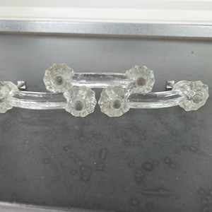 May include: Two clear glass drawer pulls featuring a curved handle design with faceted, star-shaped ends. Each pull has a central bar connecting two circular, textured mounting points that include small metal hardware components at the base.