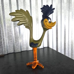 May include: A Road Runner figurine featuring a long olive green body, a large fanned tail, and a dark blue crest on its head. The character has a yellow beak, a smiling expression, and bright orange legs with black horizontal markings on the feet.