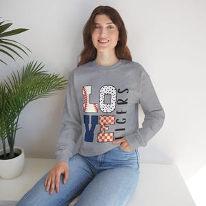 Könnte beinhalten: Ein graues Sweatshirt mit dem Wort "LOVE" in einem Baseball-Design. Das "L" ist ein Baseball, das "O" hat schwarze Punkte, das "V" ist blau und das "E" ist orange und weiß kariert. Das Wort "TIGERS" ist vertikal gedruckt.