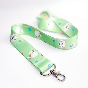 Puede incluir: Un cordón verde con un diseño de gato blanco y pequeñas flores. El cordón tiene un clip de metal plateado.