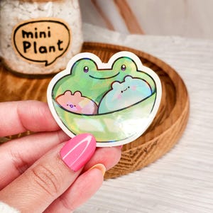 May include: A holographic sticker featuring a green frog with three smaller frogs inside. The sticker has a white outline and is on a wooden surface.