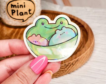 May include: A holographic sticker featuring a green frog with three smaller frogs inside. The sticker has a white outline and is on a wooden surface.