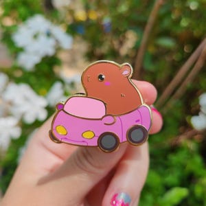 May include: A brown hippo enamel pin with a pink car design. The pin has a gold outline, black wheels, and yellow headlights. The hippo has pink cheeks and is smiling. The pin is a cute and whimsical accessory.