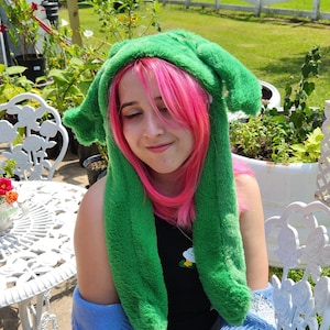 Puede incluir: Un sombrero verde brillante y afelpado con orejas largas y caídas. El sombrero lo lleva una persona con cabello rosado, que viste un top negro y un cárdigan azul. El fondo incluye una valla blanca, hierba verde y muebles de jardín.