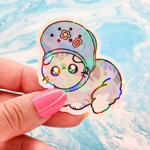 May include: A holographic sticker of a cartoon cat wearing a blue helmet with stars and a red apple. The cat has big eyes and a cute expression.