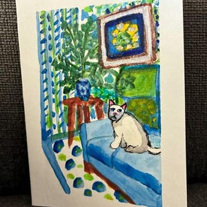 Può includere: Un acquerello su cartoncino bianco raffigurante un gatto di colore chiaro seduto su un divano blu. La scena include un tavolino marrone con un vaso blu, una pianta verde, un'opera d'arte quadrata incorniciata e tende blu a motivi geometrici.