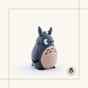 May include: A dark gray Totoro keychain with a light tan belly and white eyes. The keychain has a silver metal ring for attaching to keys or a bag. The figure is a popular character from Japanese animation.