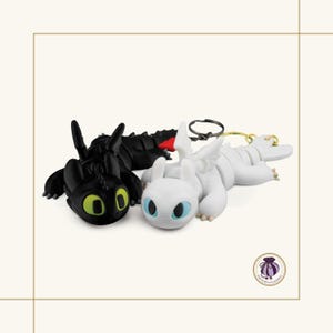 May include: Two dragon keychains featuring one black dragon with bright green eyes and one white dragon with light blue eyes. Both figures have articulated bodies, small wings, and attached metal key rings. A small circular logo with a purple bag icon is visible in the bottom right corner.