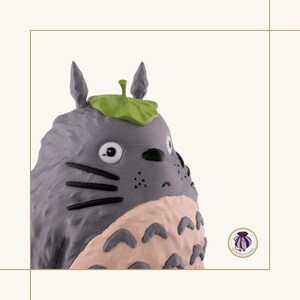 K&ouml;nnte beinhalten: Graue Totoro-Figur mit einem gr&uuml;nen Blatt auf dem Kopf. Die Figur hat einen hellrosa Bauch und schwarze Schnurrhaare. Die Figur stammt aus dem Zeichentrickfilm Mein Nachbar Totoro.