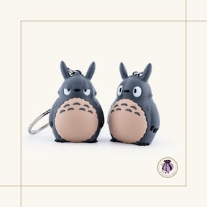 May include: Two gray Totoro keychains with tan bellies and small black eyes. Each keychain has a silver keyring attached. The keychains are standing side by side on a white background.