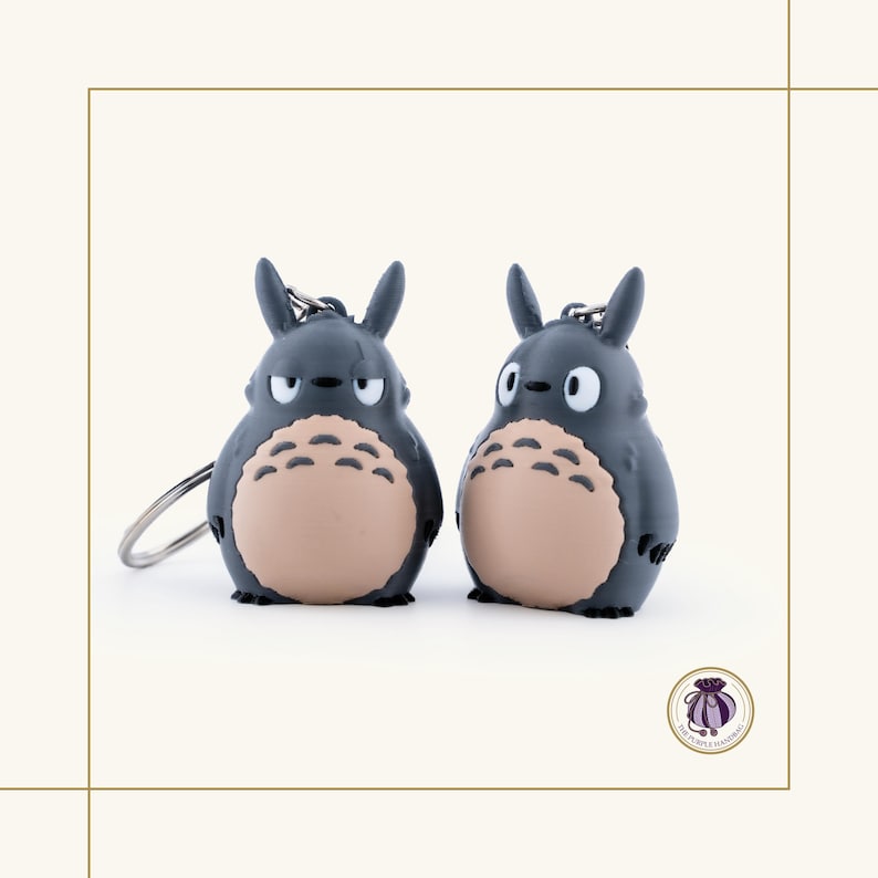 May include: Two gray Totoro keychains with tan bellies and small black eyes. Each keychain has a silver keyring attached. The keychains are standing side by side on a white background.