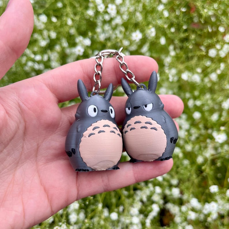 May include: A pair of gray Totoro keychains with a silver chain. Each keychain features a Totoro character with a light tan belly, black eyes, and pointed ears. The keychains are held against a background of green foliage.