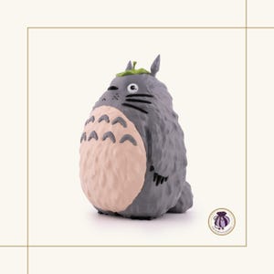 K&ouml;nnte beinhalten: Graue Totoro-Figur mit hellrosa Bauch, schwarzen Schnurrhaaren und einem gr&uuml;nen Blatt auf dem Kopf. Die Figur hat eine strukturierte Oberfl&auml;che und steht vor einem neutralen Hintergrund.