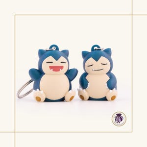 May include: Two blue and cream-coloured Snorlax keyrings. One keyring has its arms open with a smiling red mouth, and the other has its eyes closed. Both keyrings have a silver keyring attached.