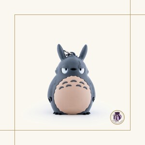 May include: A gray Totoro keychain with a light tan belly and a grumpy expression. The keychain has a silver metal ring attached to the top. The figure is a cartoon character from the animated film My Neighbor Totoro.