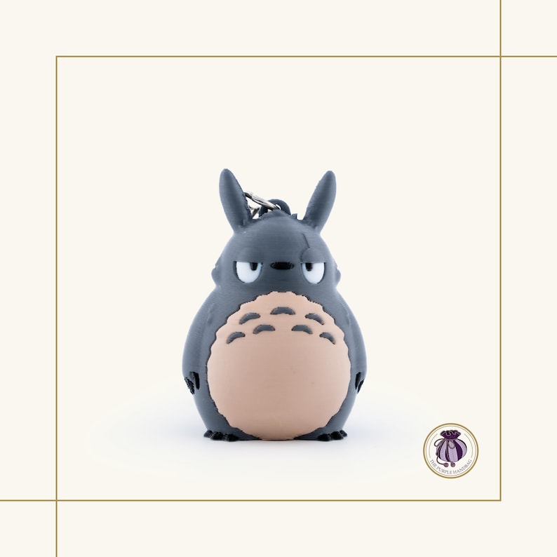 May include: A gray Totoro keychain with a light tan belly and a grumpy expression. The keychain has a silver metal ring attached to the top. The figure is a cartoon character from the animated film My Neighbor Totoro.