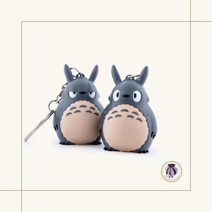 May include: Two gray Totoro keychains with beige bellies and black eyes. Each keychain has a silver metal chain and ring. The keychains are standing side by side on a white background.