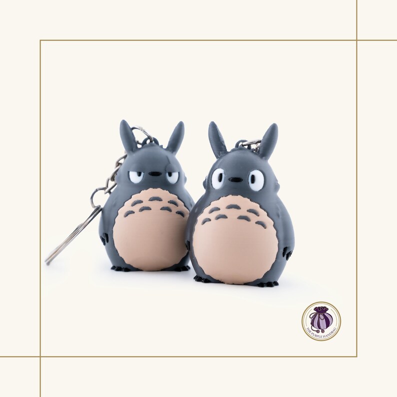 May include: Two gray Totoro keychains with beige bellies and black eyes. Each keychain has a silver metal chain and ring. The keychains are standing side by side on a white background.