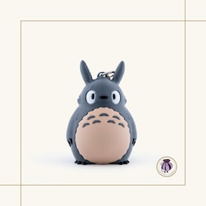 May include: A gray Totoro keychain with a light tan belly and black accents. The keychain features a silver metal clasp and a small, round, purple logo in the bottom right corner. The design is based on the animated film My Neighbor Totoro.