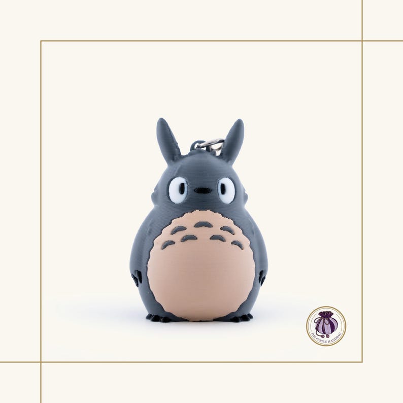 May include: A gray Totoro keychain with a light tan belly and black accents. The keychain features a silver metal clasp and a small, round, purple logo in the bottom right corner. The design is based on the animated film My Neighbor Totoro.