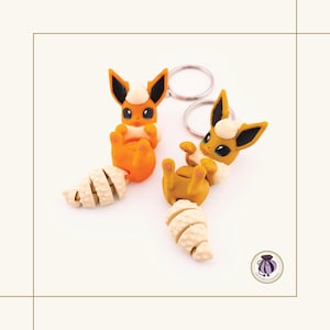 May include: Two orange and white Eevee keychains with silver keyrings, lying on their backs with a croissant. The keychains are handmade and feature black-tipped ears and blue eyes. The keychains are a unique accessory.