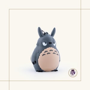 May include: A gray Totoro keychain with a light tan belly and a silver keyring. The keychain features the iconic character's distinctive features, including pointed ears and a grumpy expression. The product is a collectible item.