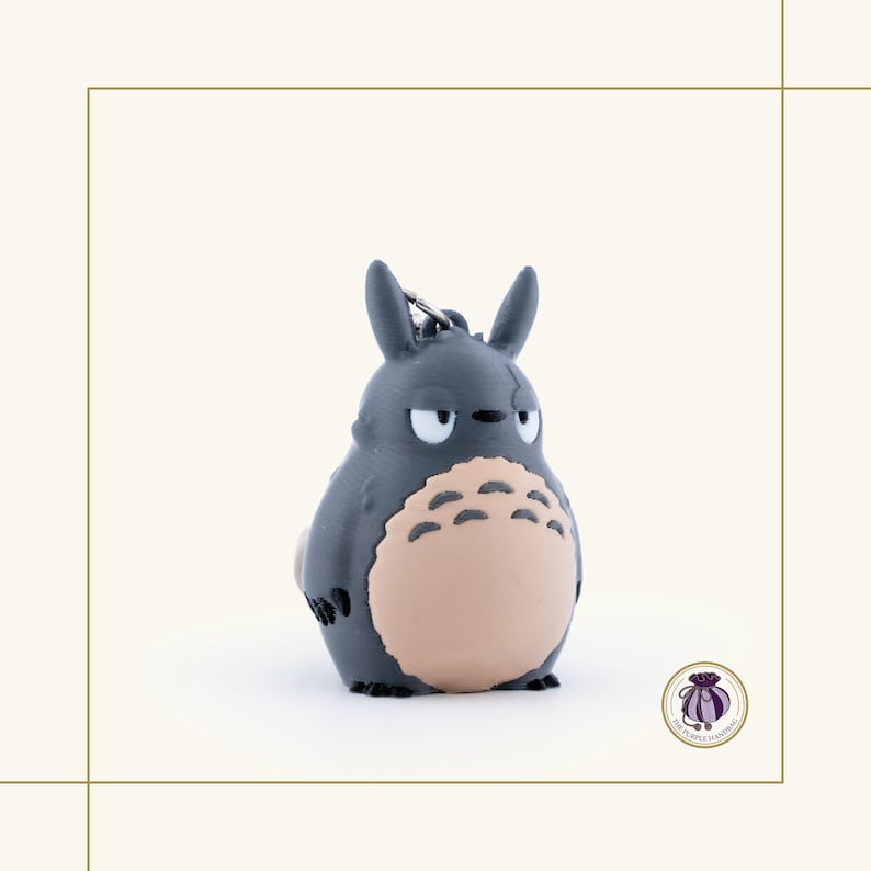 May include: A gray Totoro keychain with a light tan belly and a silver keyring. The keychain features the iconic character's distinctive features, including pointed ears and a grumpy expression. The product is a collectible item.