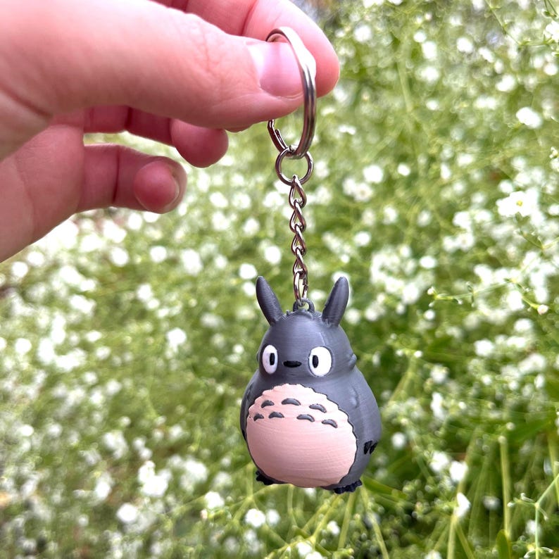 May include: A gray Totoro keychain with a silver keyring and chain. The keychain features a detailed, three-dimensional design of the character with a pink belly and white eyes. The background is a soft focus of green foliage and white flowers.