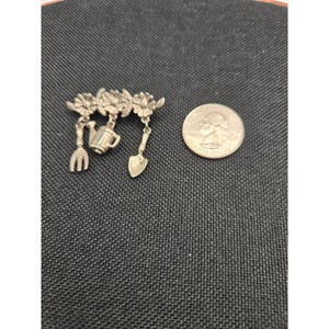Può includere: Una spilla in metallo color argento con tre attrezzi da giardinaggio pendenti: una forchetta, un annaffiatoio e una paletta, sospesi da una barra floreale. Una moneta da venticinque centesimi americana è posta accanto per dare la scala.