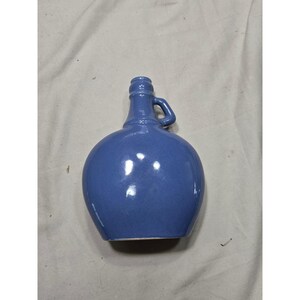 May include: A blue ceramic jug with a rounded, bulbous body and a narrow neck. The vessel features a single small handle attached to the side of the neck and a smooth, glossy finish that reflects light across its curved surface.