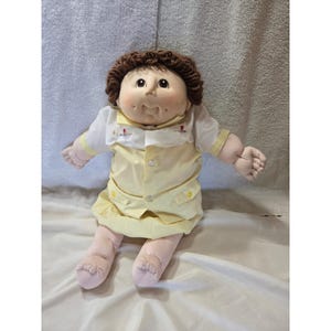 Könnte beinhalten: Eine Vintage Cabbage Patch Kid Puppe mit braunen Locken und rosigen Wangen. Die Puppe trägt ein gelbes Kleid mit einem weißen Kragenhemd und einem passenden gelben Rock. Die Puppe sitzt mit ausgestreckten Armen.