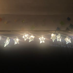 May include: A string of star-shaped lights featuring translucent, faceted covers that emit a warm glow. The lights are arranged in a horizontal line against a dark background, with the text ANGEL the MUSE visible in the lower right corner.