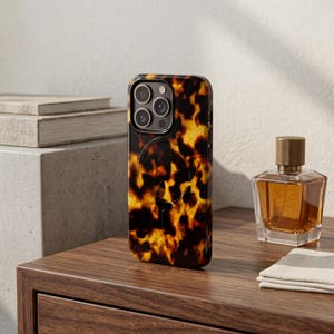 May include: A tortoiseshell-patterned phone case with a black border. The case has a glossy finish and features a camera cutout for multiple lenses. The phone case is displayed on a wooden surface next to a perfume bottle.