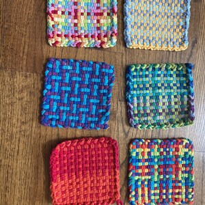 May include: A collection of six colorful, woven square potholders. Each potholder features a unique pattern and color scheme, including rainbow, blue, yellow, and red hues. The potholders are arranged on a wooden surface.