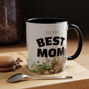 Puede incluir: Taza de cerámica blanca con interior y asa negros. La taza presenta el texto "TO THE BEST MOM EVER" en letras negras en negrita. La taza está decorada con una escena de la naturaleza con setas y plantas.