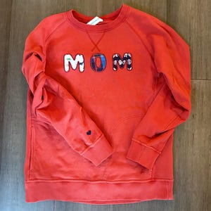 May include: A coral-colored sweatshirt with the word "MOM" appliqued across the chest in a playful, multi-patterned font. The long-sleeved top features a small blue heart on the left cuff and a crew neck design. The sweatshirt is made of a soft material.