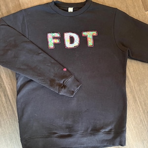 May include: Black sweatshirt with the letters "FDT" appliqued across the chest. The letters are decorated with a floral pattern in shades of pink, green, and blue. A small pink floral design is on the left sleeve.