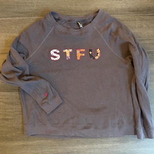 May include: A brown crew neck sweatshirt with the letters "STFU" appliqued across the chest. The letters are white with floral patterns. The left sleeve has a small pink embroidered detail.