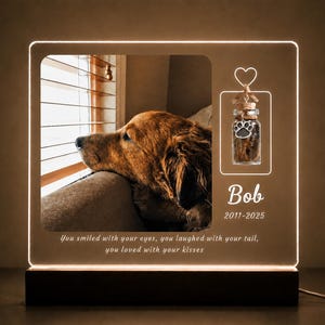 May include: A lighted memorial plaque featuring a golden retriever looking out a window. The plaque includes a small glass vial with a paw print and the name "Bob" with dates. The text reads: "You smiled with your eyes, you laughed with your tail, you loved with your kisses."