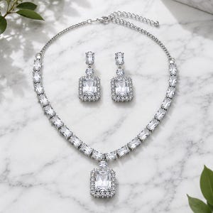 Puede incluir: Un conjunto de joyería en tono plateado que consta de un collar y un par de pendientes colgantes a juego. El collar presenta una serie de piedras rectangulares transparentes y un colgante central, mientras que los pendientes reflejan el diseño.