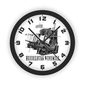 May include: A round wall clock with a black frame and a white face featuring black numbers. The center displays a black and white illustration of a Heidelberg Windmill printing press with the text HEIDELBURG WINDMILL printed below it.