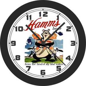 Puede incluir: Un reloj de pared redondo con marco negro y esfera blanca. El reloj presenta el texto "Hamms beer" en escritura roja sobre un oso de dibujos animados que sostiene a dos jugadores de béisbol. El reloj también tiene el texto "from the land of sky blue waters".