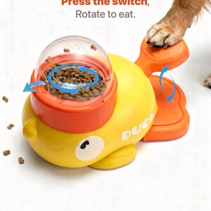 May include: A yellow and orange dog treat dispenser shaped like a duck. The dispenser has a clear top, and the text "Press the switch, Rotate to eat." is visible. A dog's paw is on the orange base.