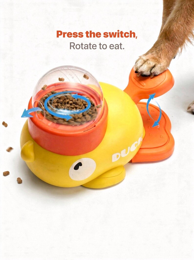 May include: A yellow and orange dog treat dispenser shaped like a duck. The dispenser has a clear top, and the text "Press the switch, Rotate to eat." is visible. A dog's paw is on the orange base.